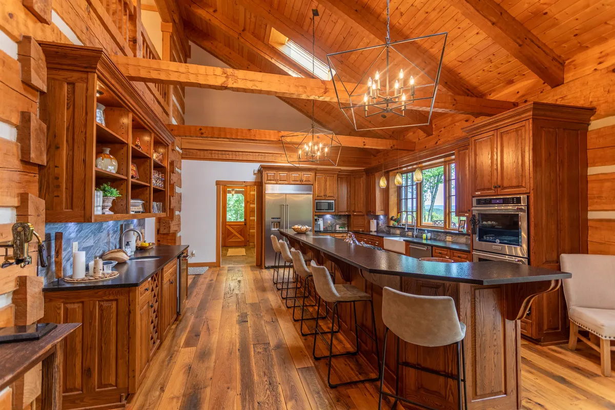 A large, inviting kitchen featuring a wooden island with bar seating, elegant chandelier lighting, open shelving displaying dishware, and expansive windows that bring in natural greenery. The space feels both cozy and functional, ideal for cooking and entertaining.