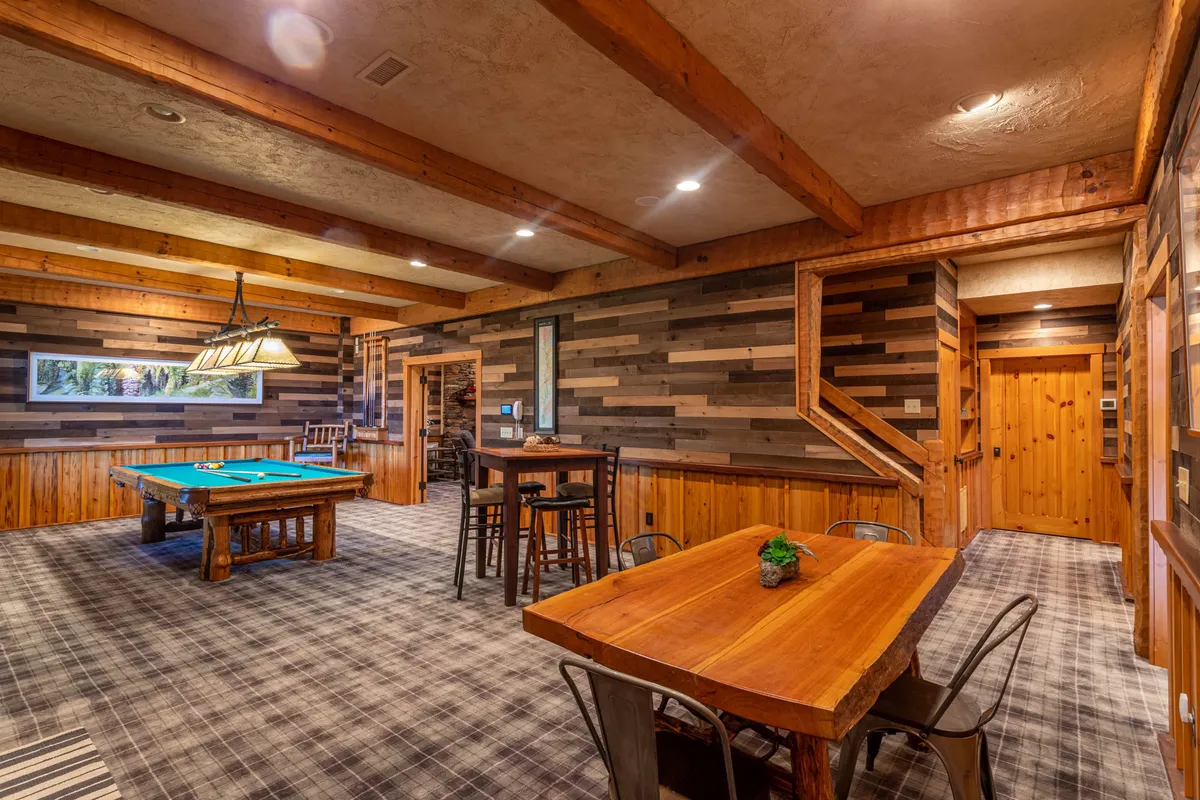 A casual entertainment space featuring a pool table, wooden beams, a dining table, and a striped accent wall. The room is designed for socializing, with comfortable seating and warm lighting, ideal for both games and casual gatherings.