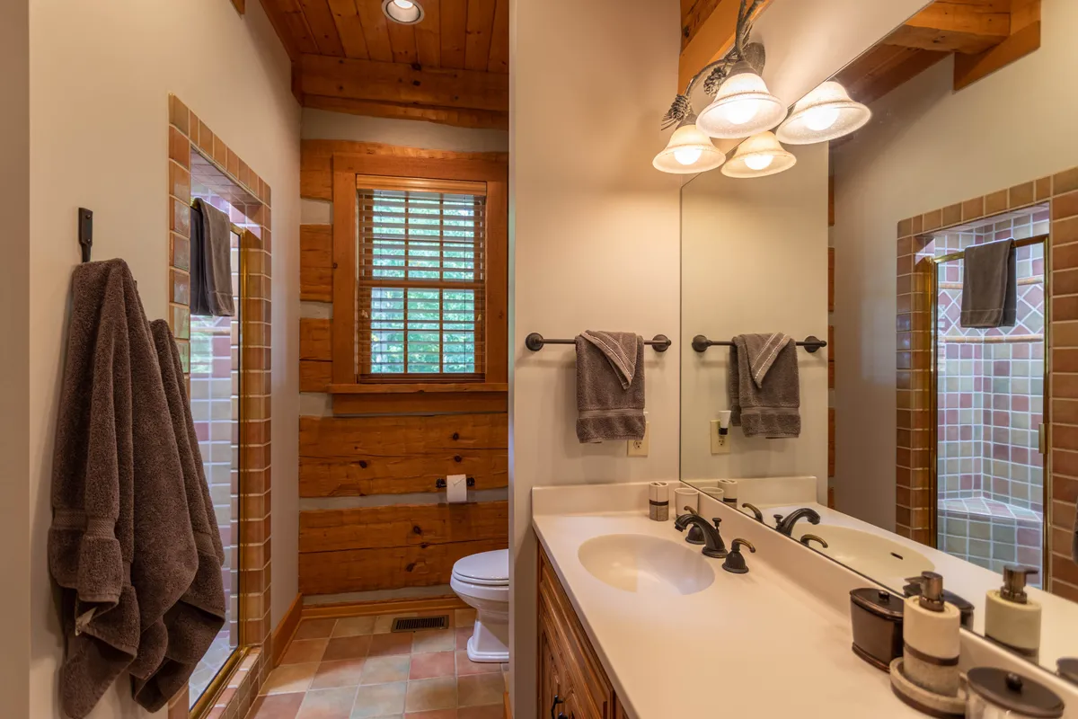 This serene bathroom features rustic wooden beams, a spacious sink area, a glass-enclosed shower, and a window with shuttered light. The overall atmosphere is calming, inviting self-care and relaxation.