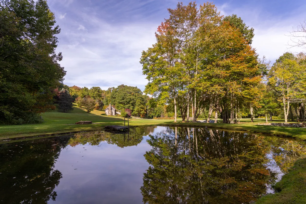 A tranquil pond surrounded by colorful trees and a distant cabin, with reflections on the still water. The landscape feels peaceful, inviting moments of relaxation, picnicking, or quiet exploration with scenic views that change with the seasons.