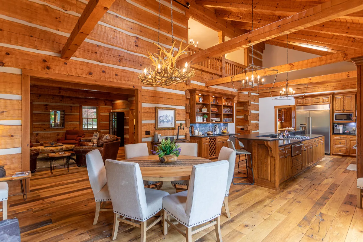A warm dining area with rustic wooden beams, a large dining table set for gatherings, a glowing chandelier, and a kitchen island visible in the background. The space exudes a sense of togetherness ideal for celebratory dinners or communal feasts.