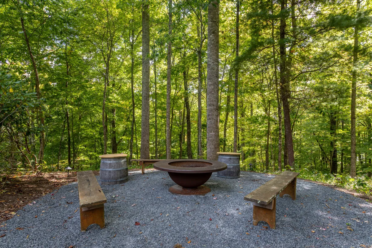 A rustic outdoor fire pit encircled by wooden benches, nestled among trees with a gravel ground underfoot. The area is clearly set up for communal gatherings, storytelling, and toasting marshmallows under the open sky.