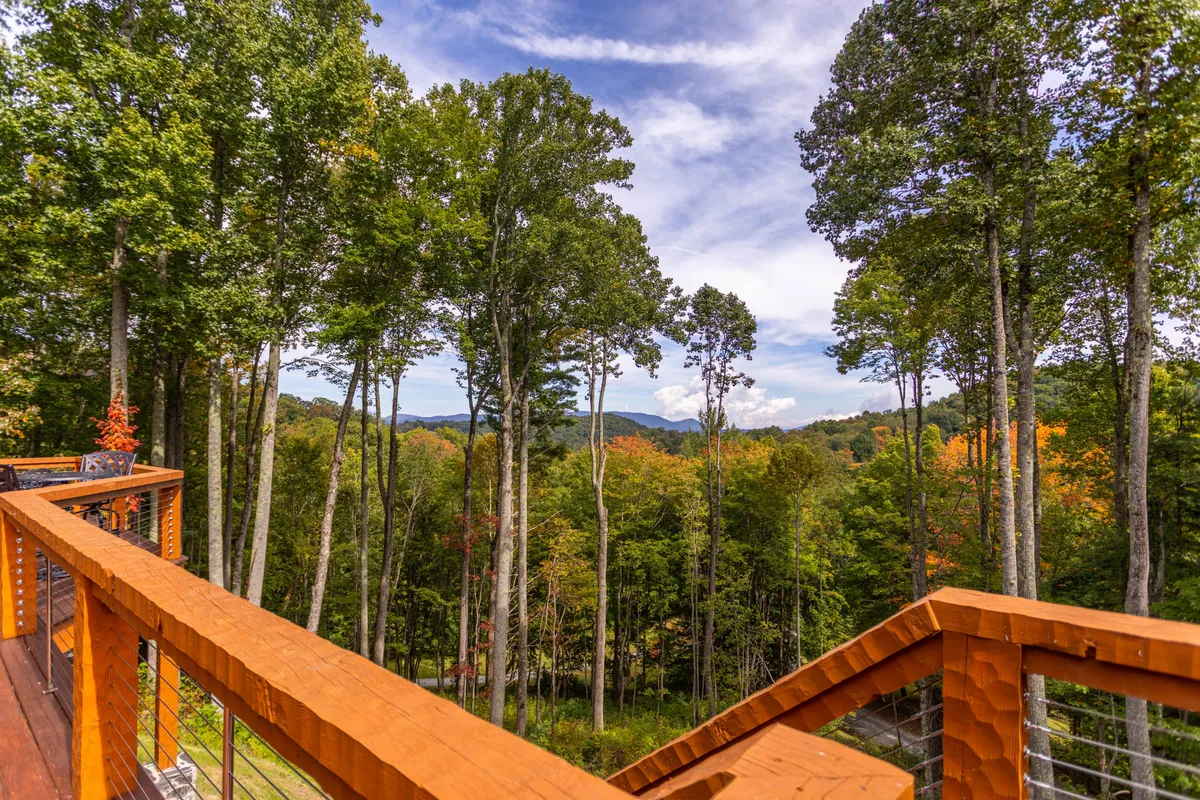 A scenic view from a deck featuring a wooden railing, overlooking a forest of lush trees with distant mountains, likely during autumn. The space invites relaxation while being immersed in panoramic nature views and the changing mountain landscape.