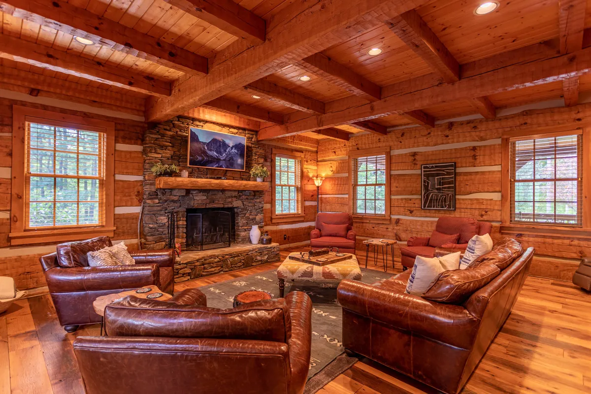 A luxurious great room with a grand stacked stone fireplace as its focal point, leather sofas arranged for conversation, exposed wooden beams overhead, striking artwork, and oversized windows revealing mountain views.