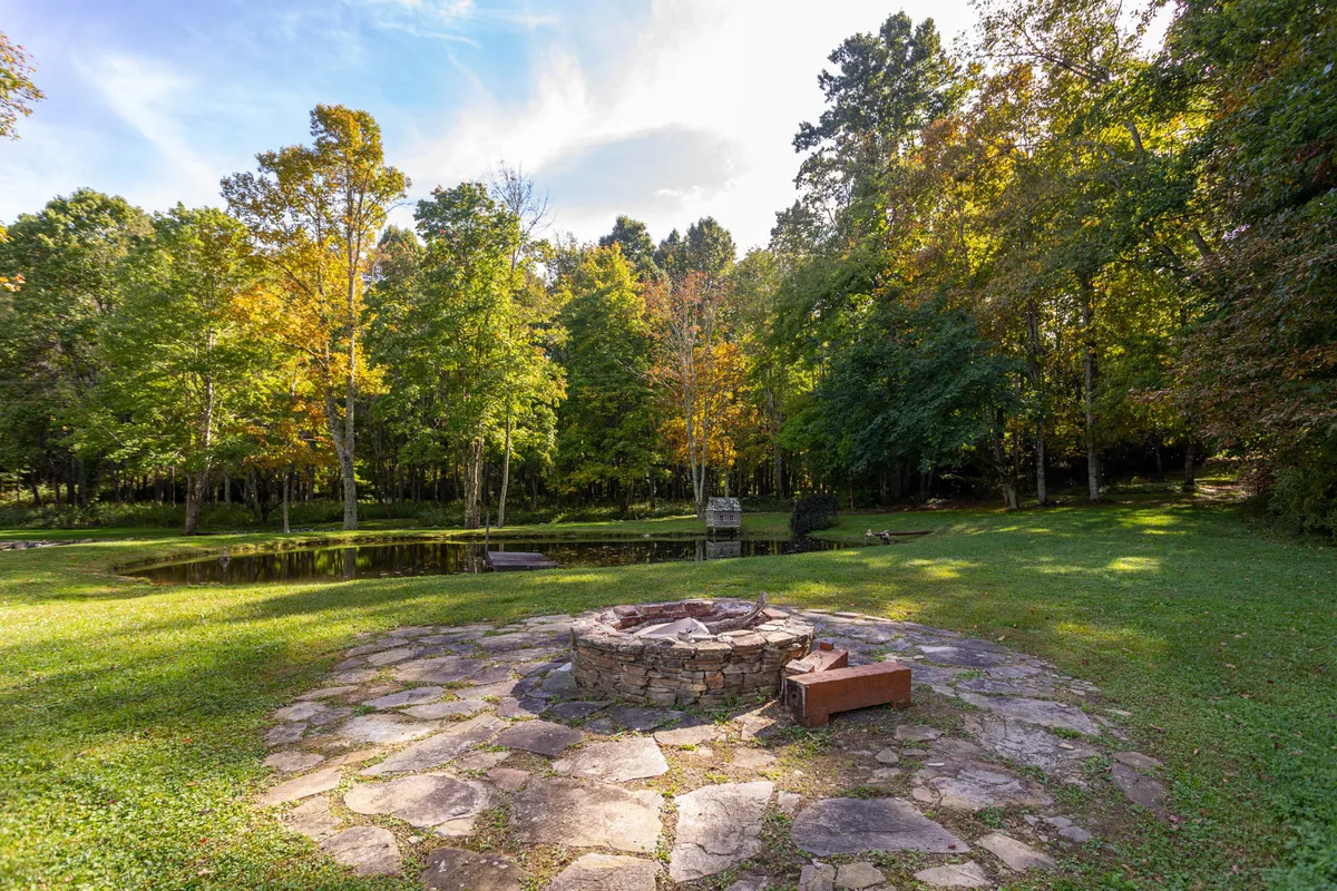 A tranquil pond sits framed by trees and open green space, with a fire pit in the foreground and ample seating for guests. The setting is perfect for daytime picnics, waterfront relaxation, or gathering with friends and family in a peaceful outdoor environment.