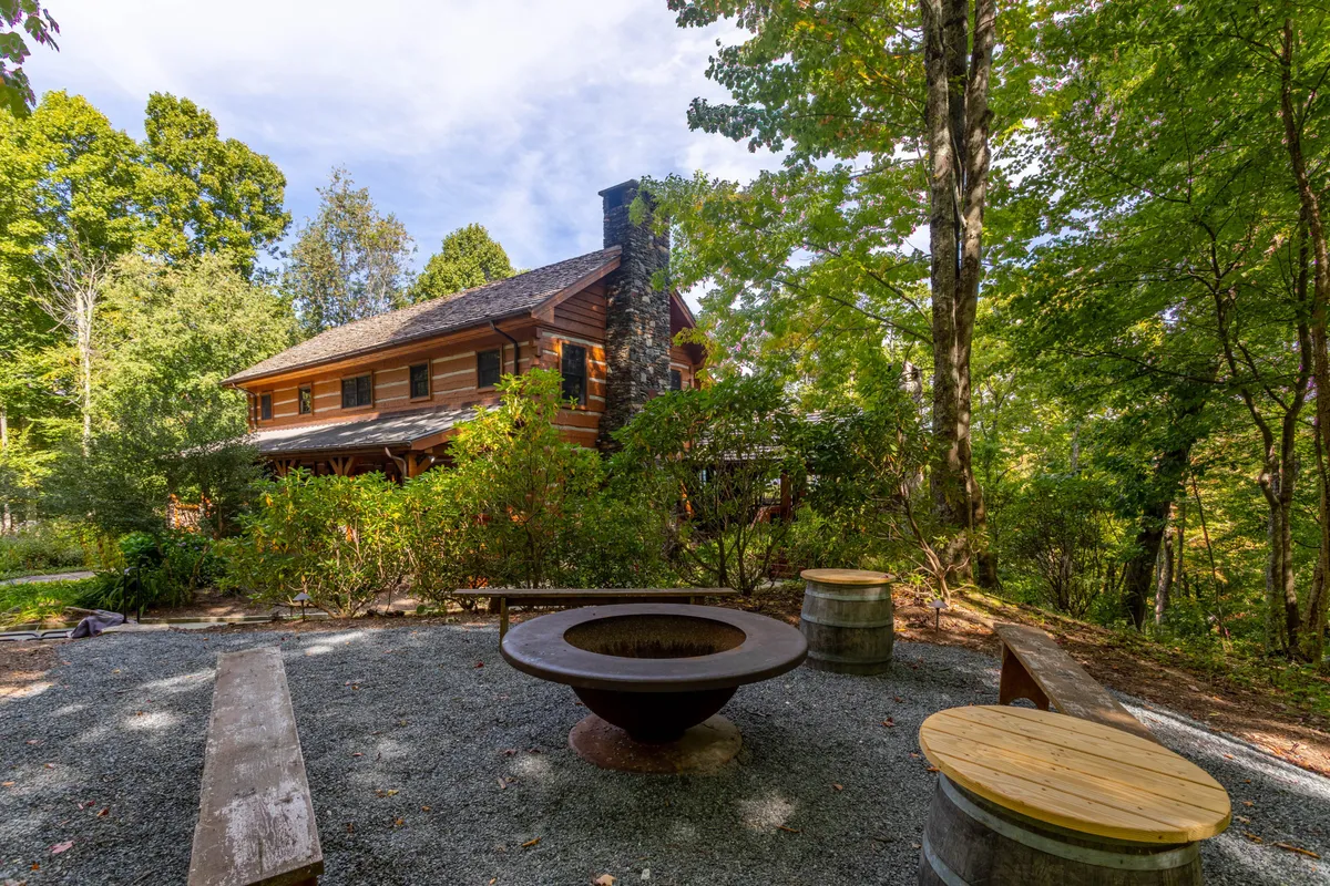 This exterior image features a log cabin set beside a fire pit, surrounded by mature trees, with a comfortable seating area gathered around the fire. The scene evokes the sense of communal gatherings and outdoor relaxation, ideal for evening storytelling and stargazing by the fire.