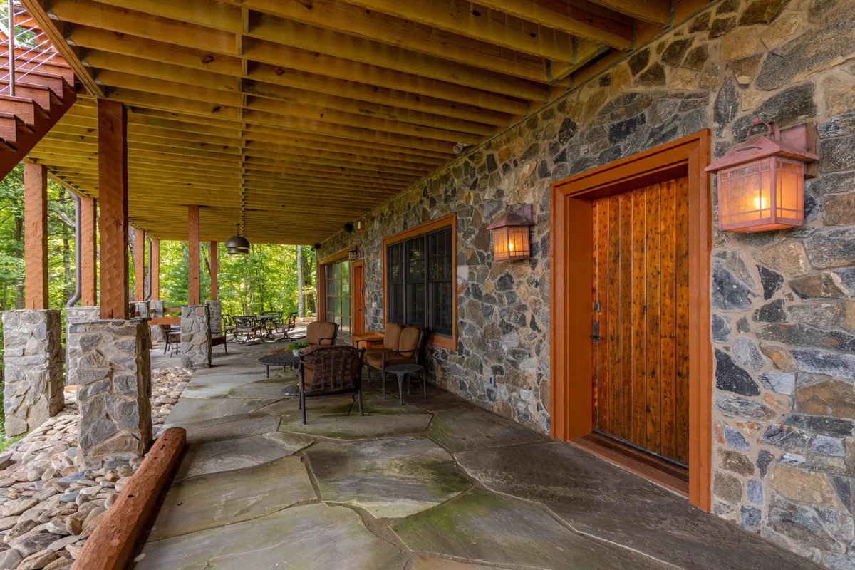 A rustic stone-faced cabin with a welcoming wooden front door, outdoor seating area, and hanging lanterns. The area is surrounded by greenery, suggesting a tranquil setting for relaxation or social gatherings just outside the main living space.
