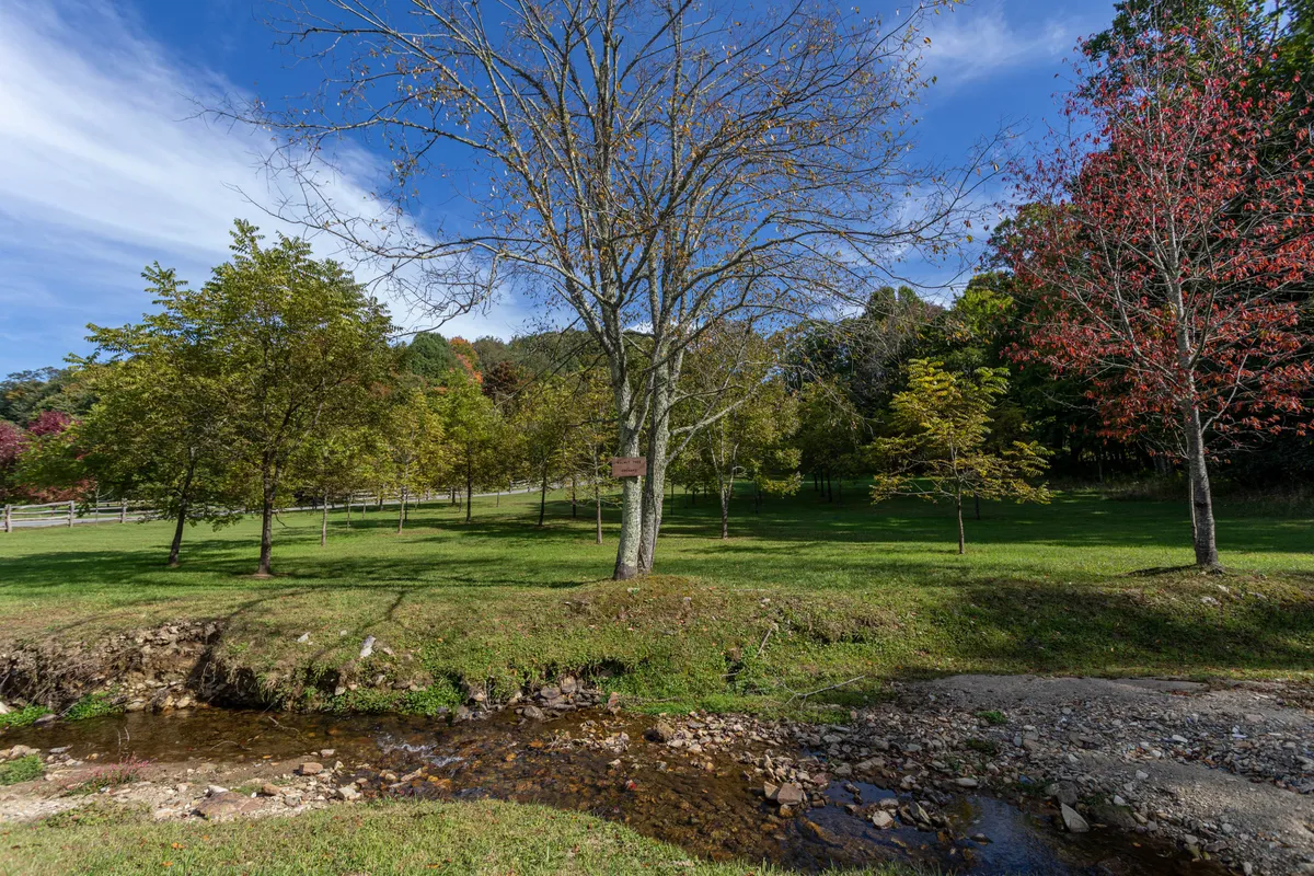 A tranquil scene featuring a gently flowing stream bordered by trees adorned in autumn foliage. A rustic wooden fence runs alongside, and the area looks perfect for leisurely picnics or peaceful walks in nature. The vibrant colors signal peak fall, while the stream and natural setting invite wildlife observation and relaxation.