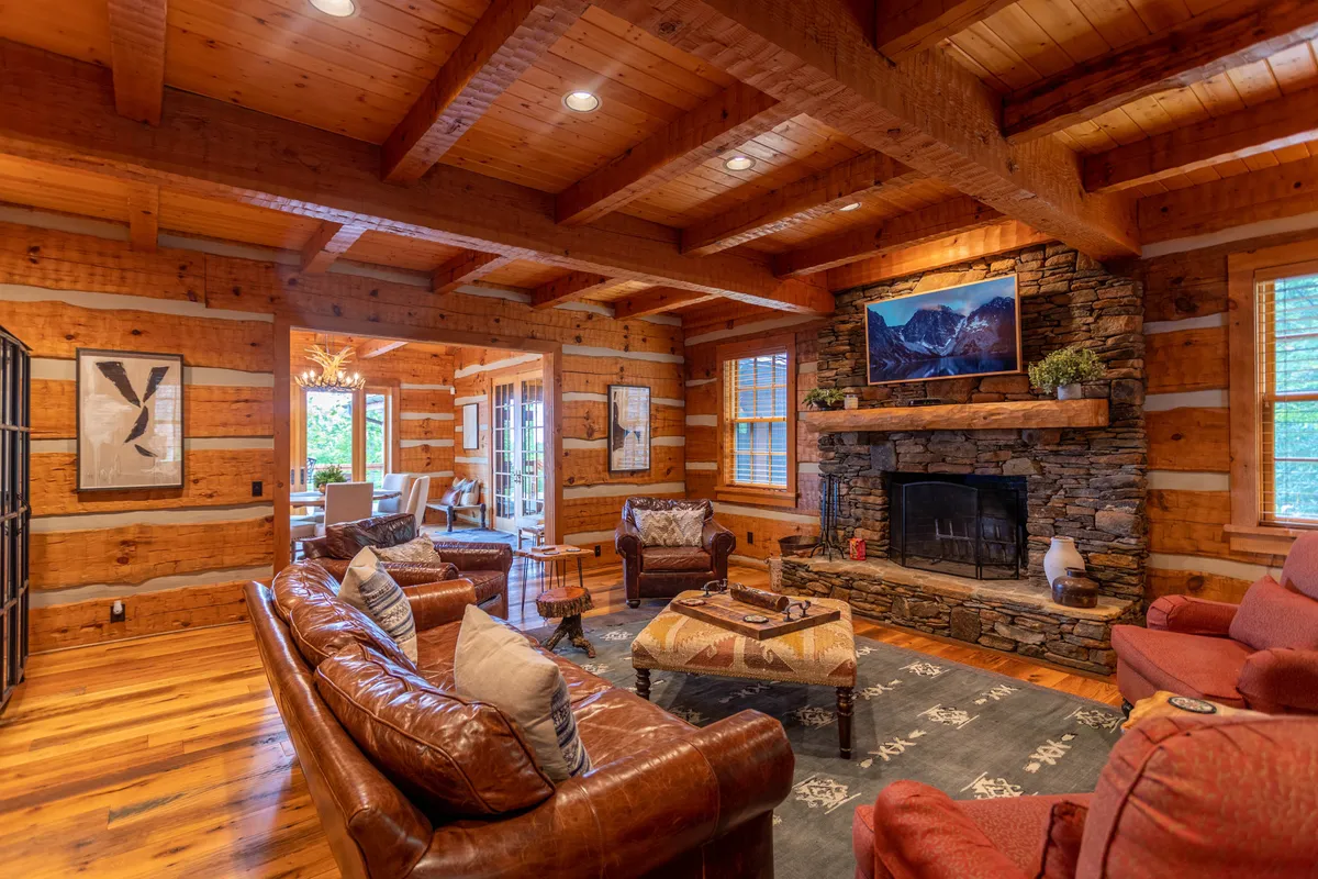A cozy screened porch featuring a grand stone fireplace, comfortable leather sofas, exposed wooden beams, and tasteful artwork. The inviting space is designed for relaxing evenings or crisp mornings in a mountain setting.